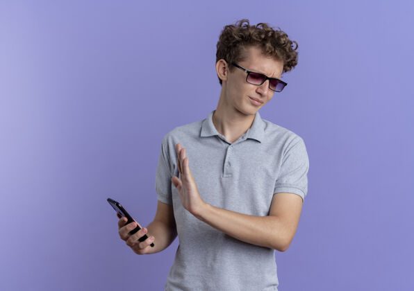 男人戴着黑眼镜的年轻人 穿着灰色马球衫 拿着智能手机 用蓝色的手做着防御的手势站着眼镜防守