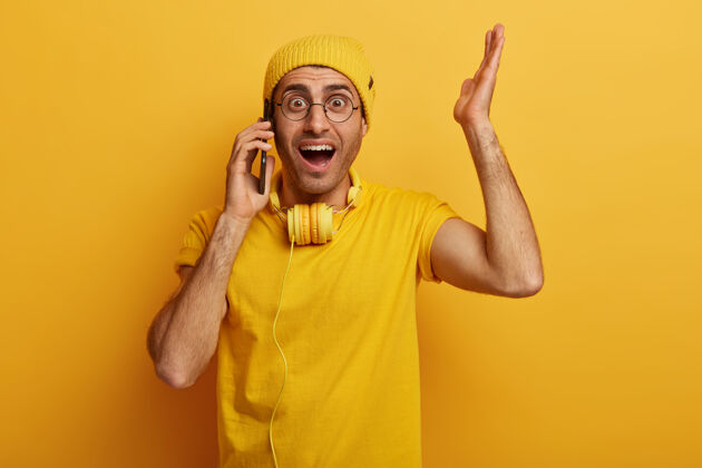 男震惊的情绪化的家伙举手 对震惊的缓和做出反应 和某人通电话 戴着时髦的黄色帽子通信帽子设备