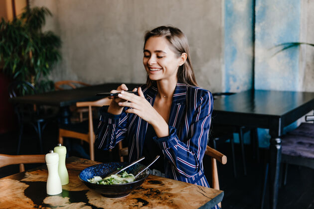 手机快乐的女孩正在为她的沙拉拍照在晚餐中微笑着的黑发女人开心的室内肖像蔬菜盘子微笑