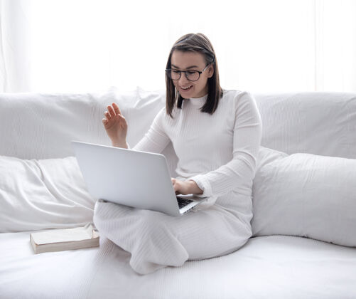 在线一个可爱的年轻女子正坐在家里的白色沙发上 穿着白色的裙子 正在用笔记本电脑工作学生学习教育