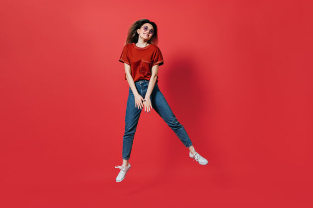 微笑穿着牛仔裤和鲜艳t恤的可爱女人在红墙上跳跃朋友年轻搞笑