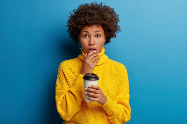 肖像照片中 一个被吓得不知所措的黑皮肤年轻女子托着下巴 在蓝色的墙上与咖啡外卖隔离个人理发情绪