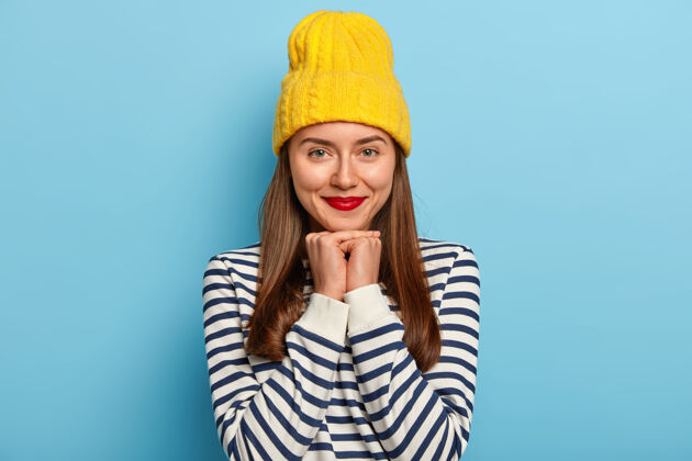 人温柔好看的黑发女人站在蓝色背景的黄色帽子和条纹毛衣 穿红色唇膏姿势乐观毛衣