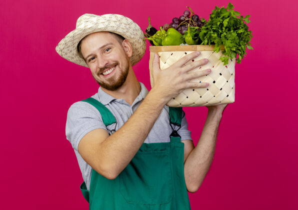 帽子面带微笑的年轻英俊的斯拉夫园丁 身着制服 戴着帽子 抱着一篮蔬菜近头望去年轻人蔬菜花园