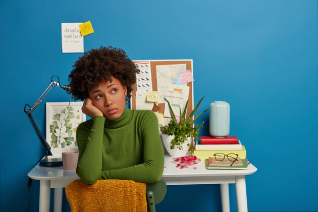 女人忧郁的黑皮肤女士看向别处 穿着绿色高领毛衣 在桌面上工作后休息 在家里蓝色背景下摆姿势桌面工作场所笔记