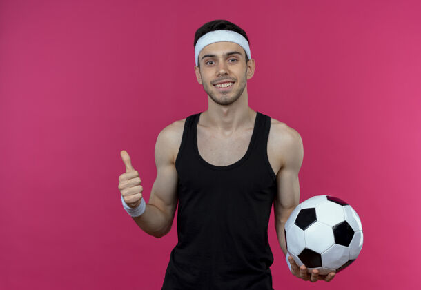 微笑戴着头巾 微笑着举着足球 竖起大拇指站在粉色墙壁上的年轻运动型男子男人拇指站着
