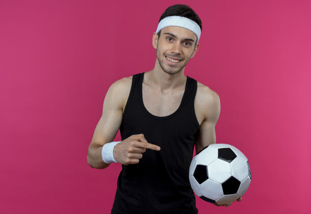 足球戴着头巾的年轻人拿着足球 用食指指着球 微笑着站在粉红色的墙上 快乐而积极目录男子手指