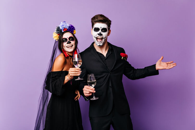 化妆舞会快乐的吸血鬼喝着紫色背景的葡萄酒一对穿着传统墨西哥僵尸服装的情侣的摄影棚照片年轻节日僵尸