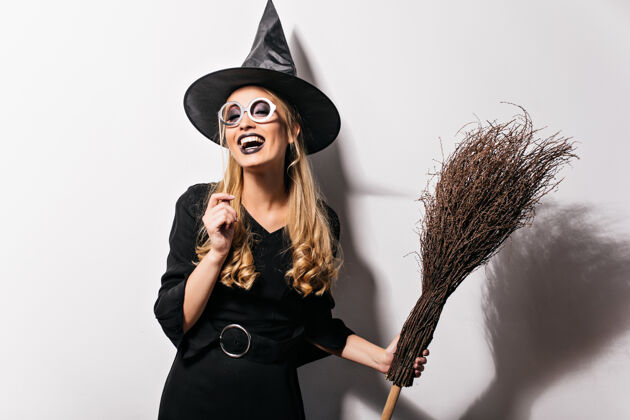 减肥戴眼镜的卷曲女巫表达万圣节的快乐穿着巫师服装拿着扫帚笑着的漂亮女孩的室内照片庆典漂亮衣服