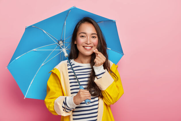 防水美丽多情的亚洲女人做韩国人一样的标志 有快乐的表情 温柔的微笑 站在伞下 穿着黄色雨衣雨秋天雨衣