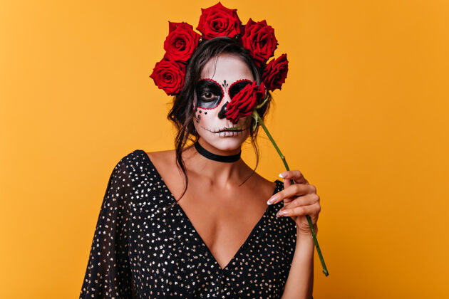 万圣节性感的墨西哥女孩拿着玫瑰站在黄色的墙上悲伤的女性僵尸在万圣节摆出鲜花的姿势传统幻想幽灵