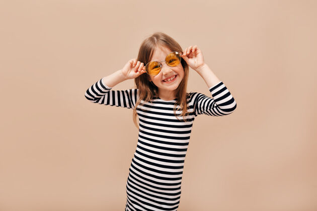 幼儿有趣快乐的6岁女孩 穿着脱衣裙 戴着橙色圆眼镜 在米色背景下带着迷人的微笑看着远方条纹微笑孩子