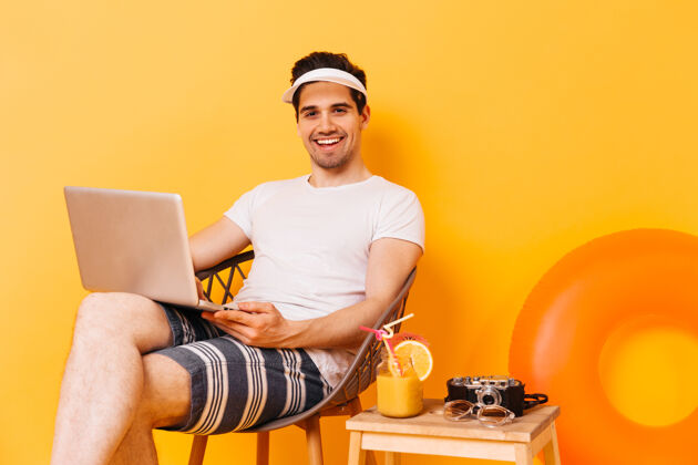 鸡尾酒戴帽子 穿白色t恤和条纹短裤的男人在笔记本电脑里工作 享受鸡尾酒包帽子短裤