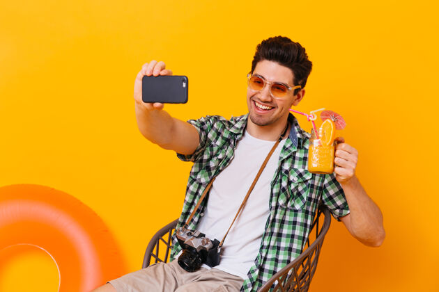 行李身穿绿色衬衫的黑发男人自拍 手里拿着橙色鸡尾酒照片中的男人拿着复古相机 在一个有充气圈的孤立空间里摆姿势男孩鸡尾酒包
