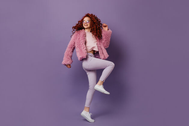 发型卷曲的红发女孩穿着白色运动鞋和裤子在紫色的空间里跳舞和笑经典魅力少女