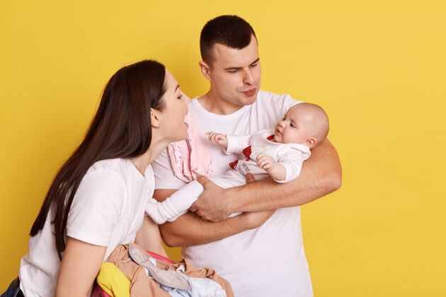 悲伤一对幸福的夫妇在黄色的背景下拥抱和看着新生的孩子 带着爱和微笑和小女儿交谈 父母穿着白色t恤 快乐的家庭在室内宝贝女性困倦
