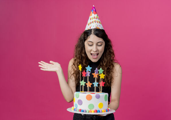 卷发年轻漂亮的女人 卷发 戴着节日帽 手里拿着生日蛋糕 站在粉色的墙上 快乐而兴奋地庆祝生日蛋糕年轻站着