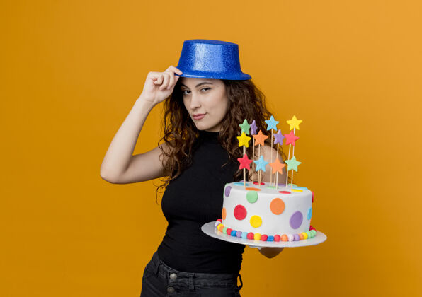 女人一个年轻漂亮的女人 卷发 戴着节日帽 手里拿着生日蛋糕 自信地站在橙色的墙上自信年轻卷发