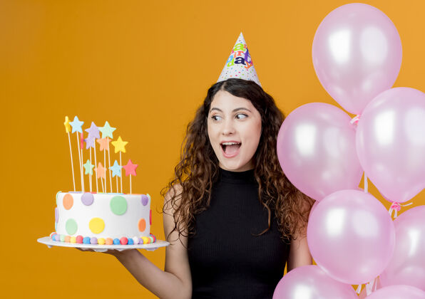 卷曲一个年轻漂亮的女人 卷发 戴着节日帽 手里拿着气球和生日蛋糕 站在橙色的墙上 快乐而兴奋地庆祝生日兴奋蛋糕站着