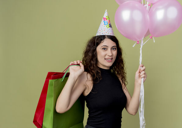 气球一位年轻漂亮的女士 卷发 戴着节日帽 手里拿着气球和生日礼物 快乐而积极地微笑着站在明亮的墙上积极抱着欢呼