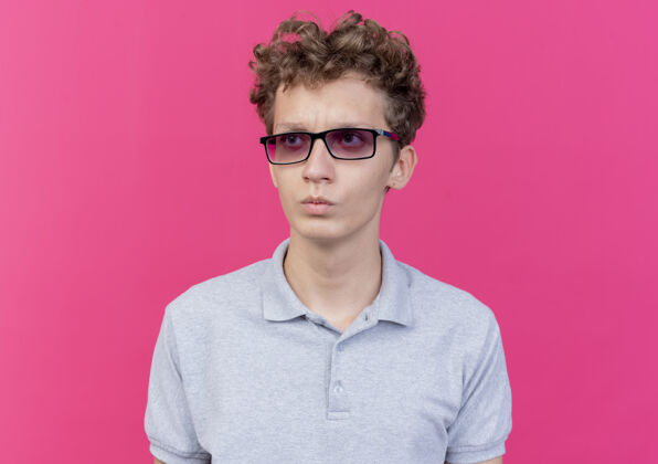 戴一个戴着黑眼镜 穿着灰色马球衫的年轻人站在粉红色的墙上 严肃地看着一边眼镜严肃靠边站