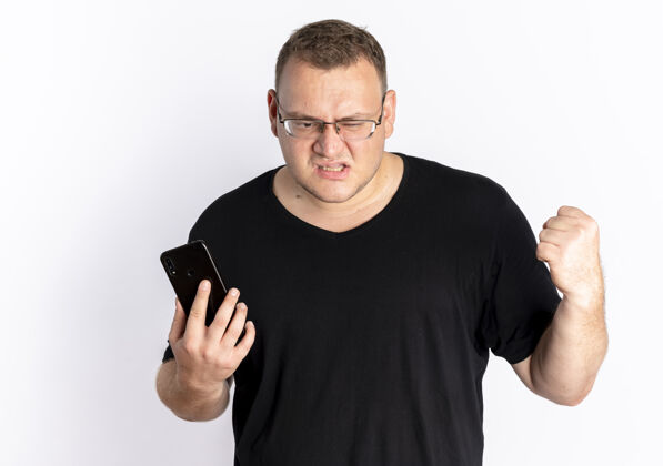 男人戴眼镜的超重男子身穿黑色t恤手持智能手机握紧拳头愤怒的脸站在白墙上戴着抱着愤怒