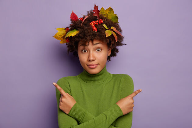 索引照片中 困惑的黑皮肤女人 头发上留着秋叶 双手交叉在胸前 侧指 身穿绿色套头衫 孤立在紫色背景上叶子怀疑显示