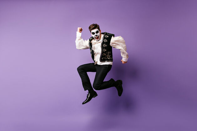 万圣节穿着万圣节服装的欢笑跳跃的男人在紫色墙壁上穿着墨西哥妆跳舞的兴奋的家伙的室内照片多彩死亡日骷髅