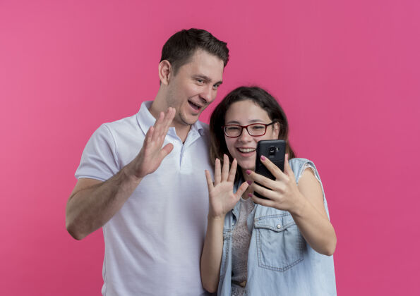 视频一对穿着休闲服的年轻夫妇站在粉红色的墙上 手上挥舞着微笑 手里拿着视频电话女人站立欢呼