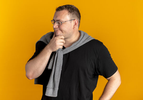 男人一个戴着眼镜 身穿黑色t恤 手放在下巴上 表情沉思的超重男子站在橘色的墙上沉思戴着眼镜