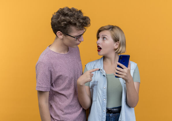 穿着一对年轻漂亮的情侣一男一女穿着休闲服男人拿着智能手机站在橘红色的墙上和女朋友吵架休闲男人手机