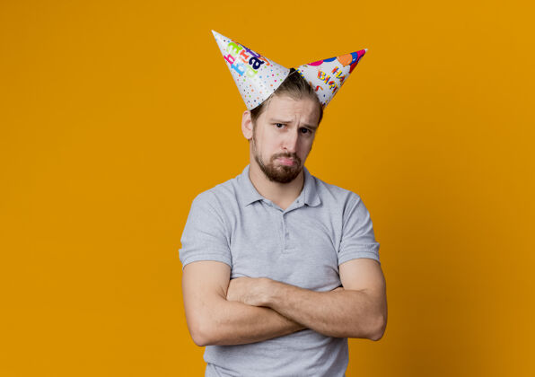 男人戴节日帽的年轻人站在橘色的墙上不高兴地庆祝生日看帽子年轻人