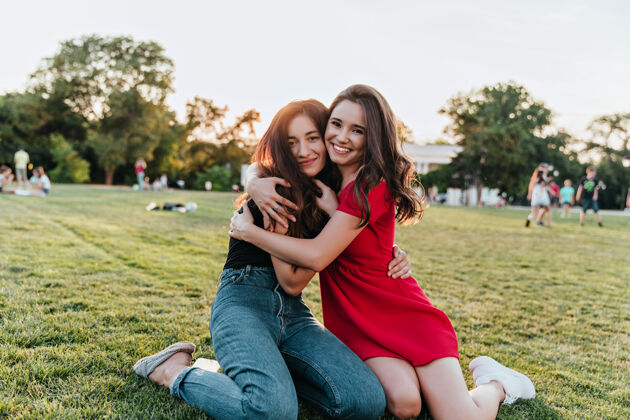户外穿着牛仔裤的害羞女人坐在草地上拥抱她的朋友幽默的女人在公园里享受夏日周末积极美丽年轻