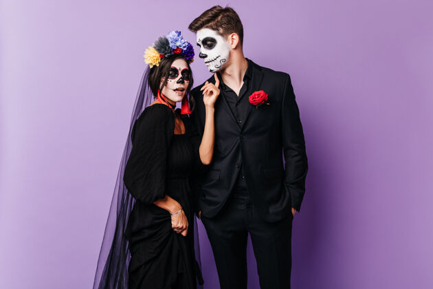 死亡可爱的僵尸女孩戴着五颜六色的花环和男朋友站在一起一对身着muertos服装的高加索夫妇在紫色背景的万圣节摆姿势西班牙幻想五颜六色