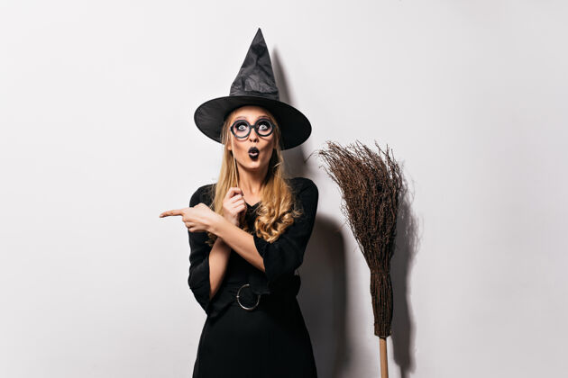 万圣节戴着眼镜的巫师站在白色的墙上滑稽的情绪巫师拿着帽子和扫帚摆姿势魔术师幽灵魔鬼