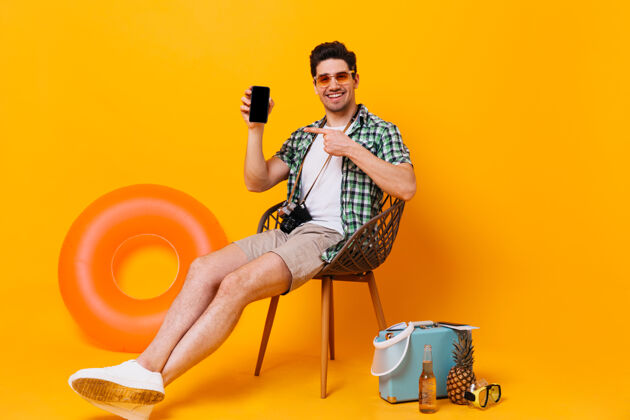 背包一个乐观的男人穿着绿色衬衫和米色短裤 在他的智能手机上展示一个戴着太阳镜的家伙坐在椅子上 拿着手提箱 啤酒 充气圈男孩啤酒行李
