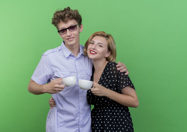 在一起年轻漂亮的夫妇站在一起 捧着咖啡杯 微笑着 在绿色的墙上 幸福地相爱年轻抱着站着