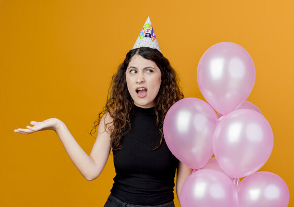 气球一个年轻漂亮的女人 卷发 戴着节日帽 手里拿着气球 站在橘黄色的墙上 看起来很困惑生日派对的概念抱着帽子困惑