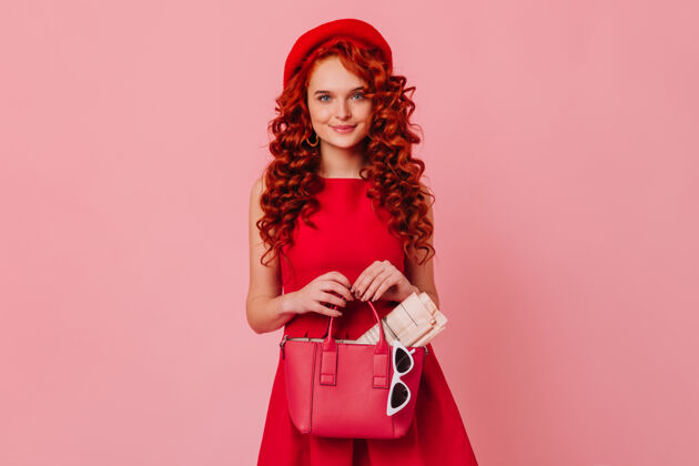 女主角长发可爱的女士 戴着红帽子 穿着鲜艳的裙子 在粉红色的空间里摆着姿势蓝色眼睛的女士拿着皮包 杂志和眼镜头发浪漫女性