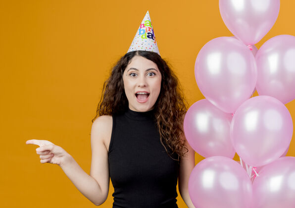 手指一位年轻漂亮的女士 一头卷发 戴着节日帽 手里拿着气球 身材匀称 兴奋地用手指着站在橘色墙上的生日派对概念拿着指着兴奋