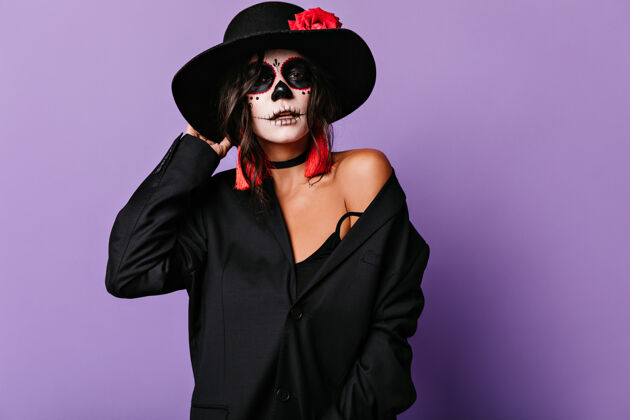 面纱穿着超大号夹克 戴着万圣节面具的时髦女人摸着帽沿紫丁香墙上有一个漂亮的晒黑女孩的画像拉丁语嘉年华死亡