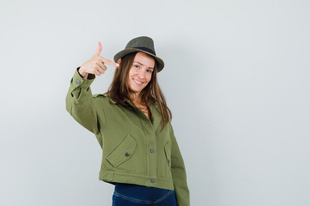 手指年轻女性在夹克 裤子 帽子上做手指标志 看起来很自信正面图微笑年轻正面