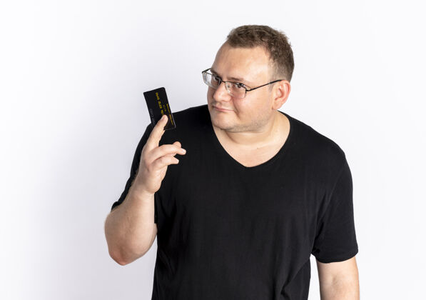 超重一个戴眼镜 身穿黑色t恤 出示信用卡的超重男子站在白色的墙上困惑地看着摄像机困惑男人穿