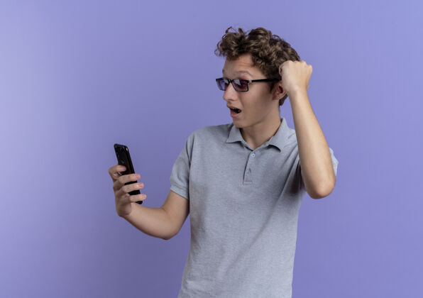年轻穿着灰色马球衫的年轻人看着他的智能手机屏幕 握紧拳头站在蓝色的墙上 快乐而兴奋马球站兴奋