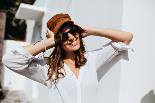 白色建筑戴着太阳镜的美女戴着灯芯绒帽子在白色的墙壁上画着兴高采烈的女孩棕榈树金发白衬衫