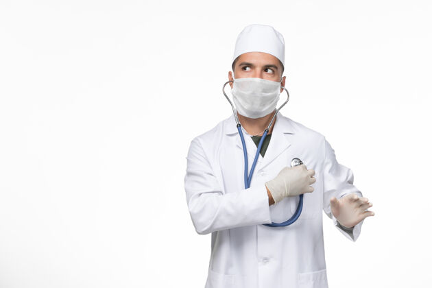 男性正面图：男医生穿着医疗服 戴着防冠状病毒口罩 在白墙上用听诊器观察冠状病毒疾病大流行正面冠状病毒大流行