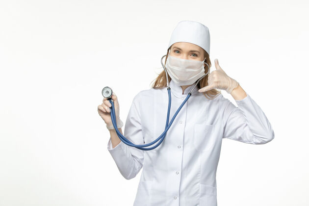 视图前视女医生身穿医疗服戴口罩因冠状病毒手持听诊器白桌病毒大流行-冠状病毒病女性冠状病毒佩戴