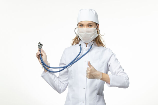 视图正面图女医生穿着白色医疗服 带无菌口罩 因冠状病毒使用听诊器在白色办公桌上大流行病毒冠状病毒预防正面套装