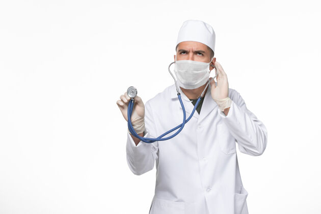 男性正面图：男医生穿着医疗服 戴着口罩 用听诊器在白色墙壁上对抗冠状病毒-疾病大流行疾病使用医生