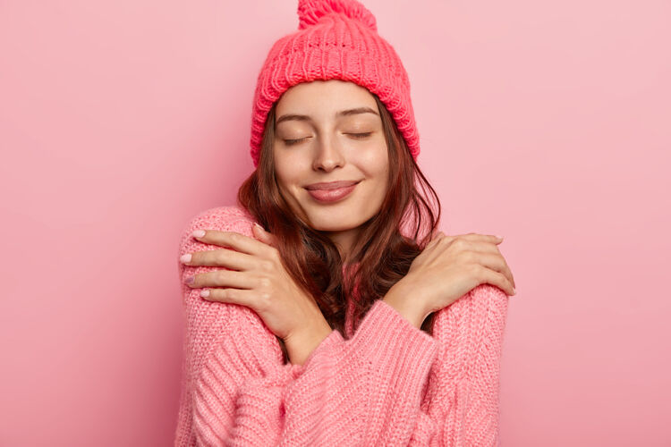 女人满意的黑发女人的肖像拥抱自己 享受舒适的针织温暖毛衣 保持眼睛紧闭 购买新的冬季服装 隔离在粉红色的背景享受年轻站